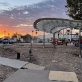 Construction site at sunrise with a partially built walkway and curved shade structure, with construction equipment and workers in the background.